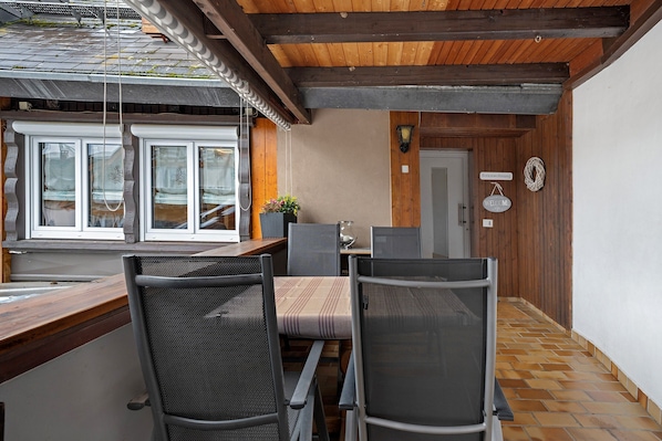 A covered outdoor dining area with a wooden ceiling, a window with blinds, and a door with a welcome sign.