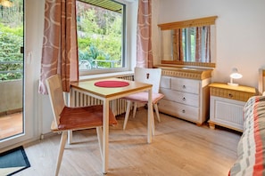 Une pièce avec un plancher en bois, une fenêtre avec des rideaux, un bureau avec une chaise, une commode, un miroir et une table de chevet avec une lampe.