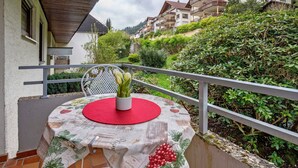 Un balcone con un tavolo ricoperto da una tovaglia di vigne, una sedia bianca e un vaso con fiori.