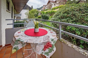 Un balcón con una mesa cubierta con mantel de parra, una silla blanca y un florero con flores.