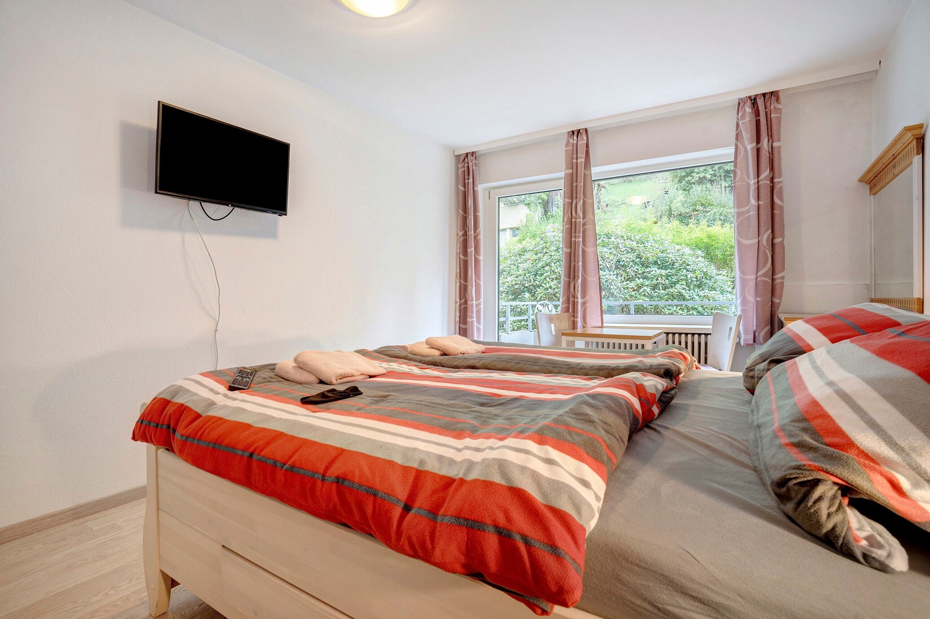 A bedroom with two beds, a TV mounted on the wall, and a window with curtains.