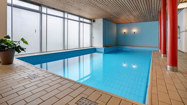 Una piscina interna con pavimento piastrellato, soffitto in legno e ampie finestre.