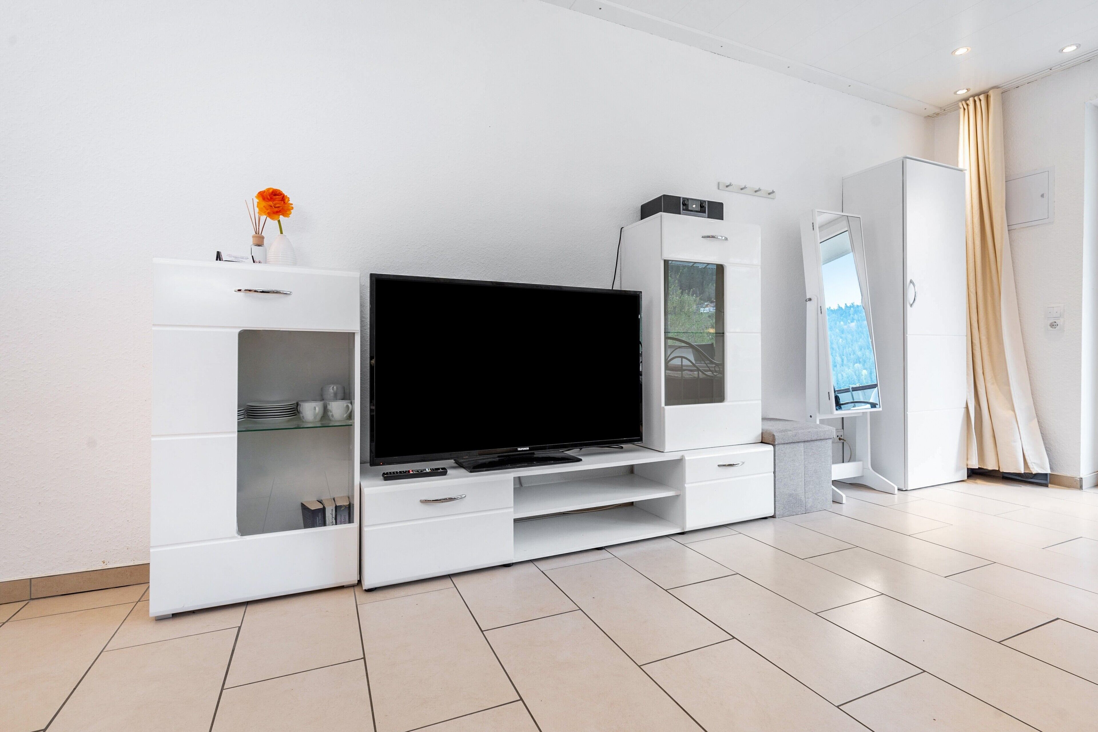 A modern living room with a flat-screen TV, a white cabinet with glass doors, and a beige curtain.