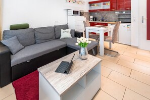 Un salon moderne avec un canapé gris, une petite table sur laquelle se trouve un ordinateur portable, un vase avec des fleurs blanches, et une cuisine en arrière-plan.