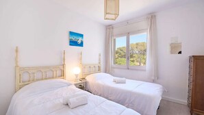 A room with two beds, a nightstand with a lamp, a wooden dresser, and a window with a view of trees. - Apartment in Calella near Sand Beach (Calella de Palafrugell, Llafranc)