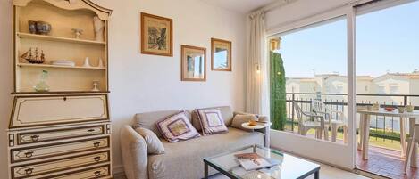 A living room with a sofa, coffee table, and a cabinet with decorative items. There are framed pictures on the wall and a balcony with outdoor furniture.