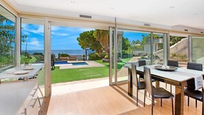 Un comedor moderno con una mesa grande y sillas, vistas a una piscina y jardín, y una pared de cristal con vistas al mar.