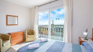 Quarto com cama, cadeira, cômoda, escrivaninha com luminária e varanda com vista.