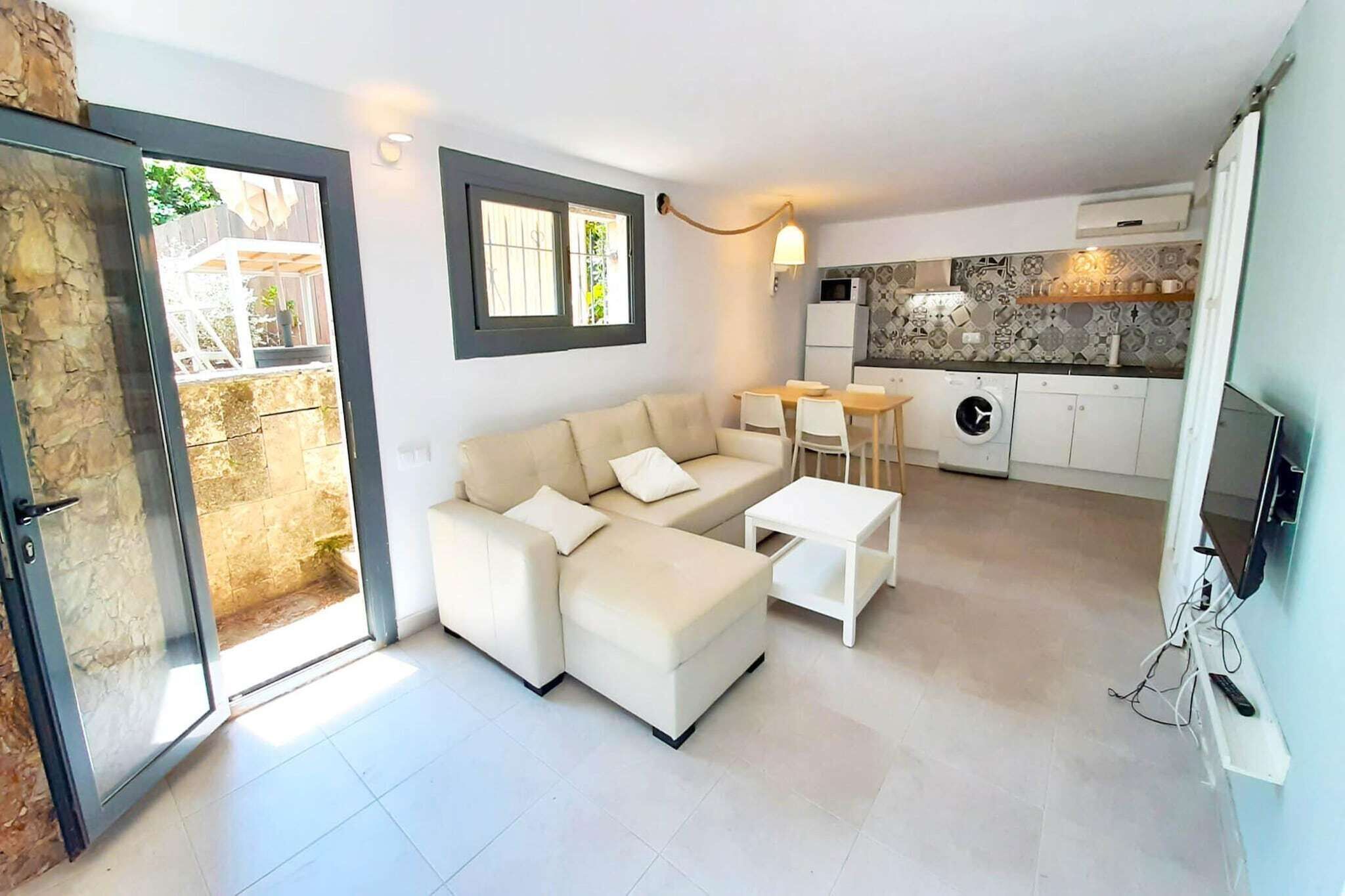 Una sala de estar moderna con un sofá, una mesa de centro y un televisor. Se observa una zona de cocina con lavadora y una mesa de comedor.