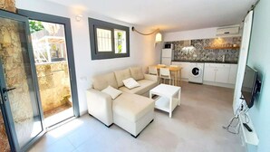 A modern living room with a sofa, a coffee table, and a TV. There is a kitchen area with a washing machine and a dining table.