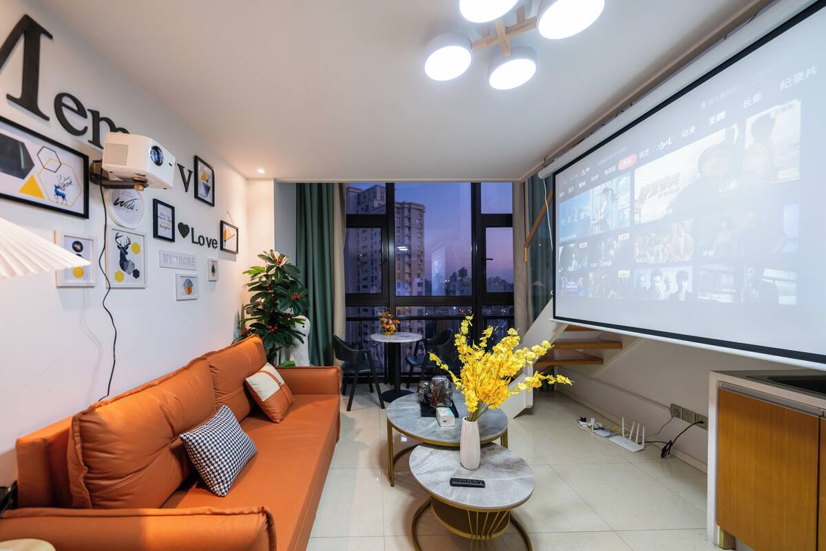 A modern living room with a large screen, orange sofa, and a small table with a vase of flowers.