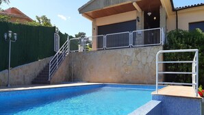 A swimming pool with a stone wall and metal railings, a staircase leading to a covered area, and a clear blue sky.