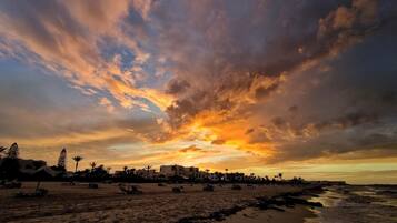 A beach with a sunset, palm trees, and buildings in the background.
