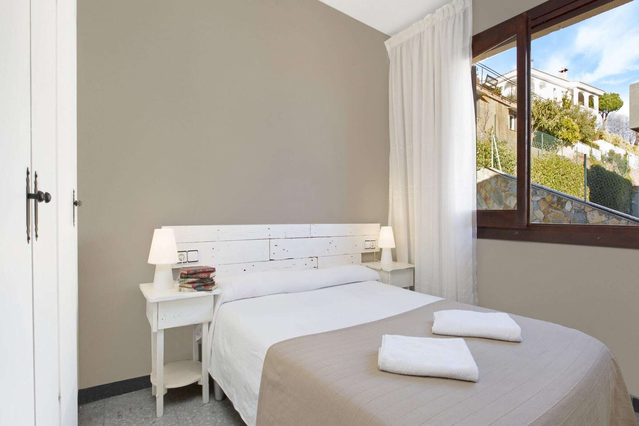 A hotel room with a bed, a bedside table with a lamp, a window with a view of buildings and trees, and a door.