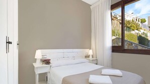 Quarto de hotel com cama, mesa de cabeceira com abajur, janela com vista para edifícios e árvores, e porta.
