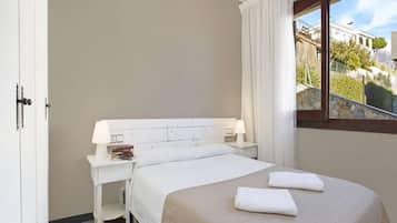 A hotel room with a bed, a bedside table with a lamp, a window with a view of buildings and trees, and a door.