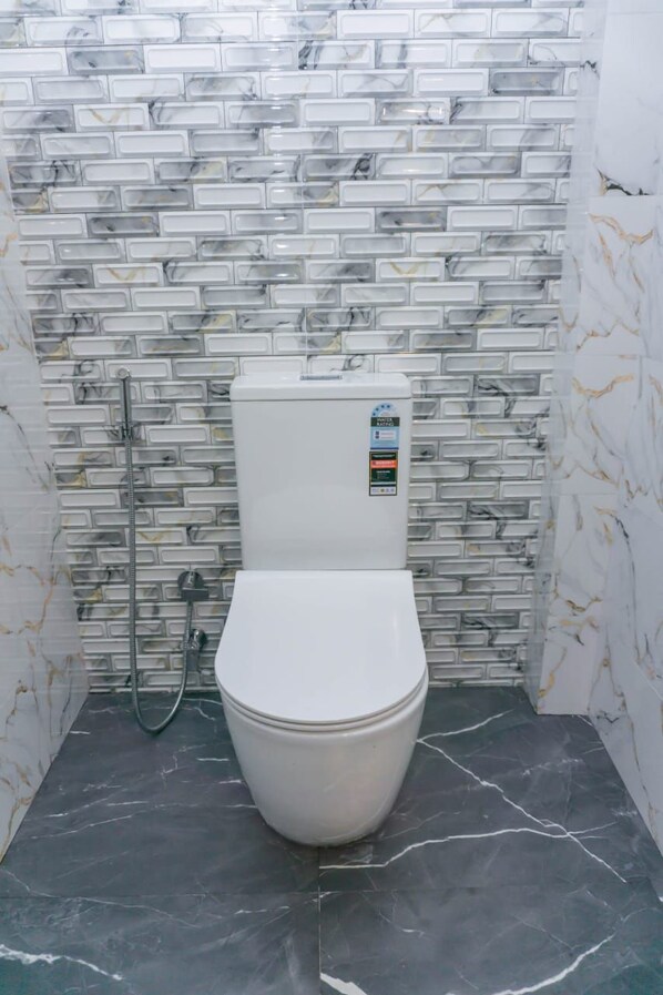 A bathroom with a white toilet, a marble-patterned wall, and a black marble floor. - Rome Transit Hotel (Negombo)