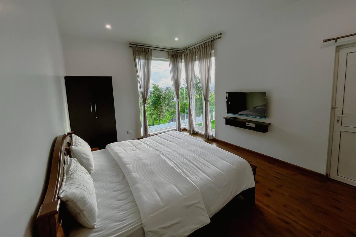 Une chambre à coucher avec un grand lit, une armoire, un téléviseur fixé au mur et une fenêtre avec des rideaux.