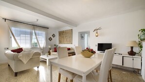 A modern living room with a white sofa, a dining table with chairs, a TV, and a lamp.