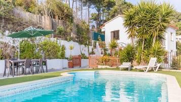 Una piscina con un deck que la rodea, un área de comedor con sillas y un edificio blanco con techo rojo.