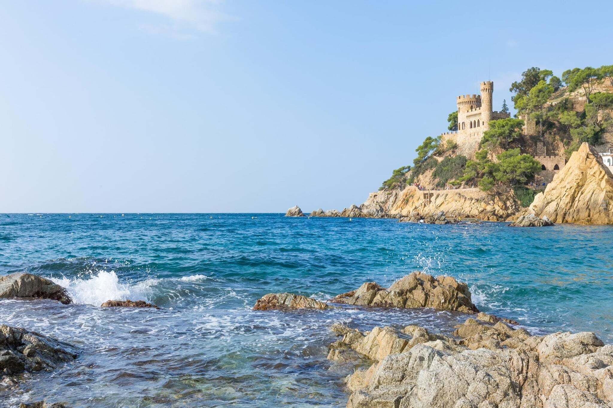 A coastal view with a castle on a cliff, clear blue sea, and rocky shoreline.