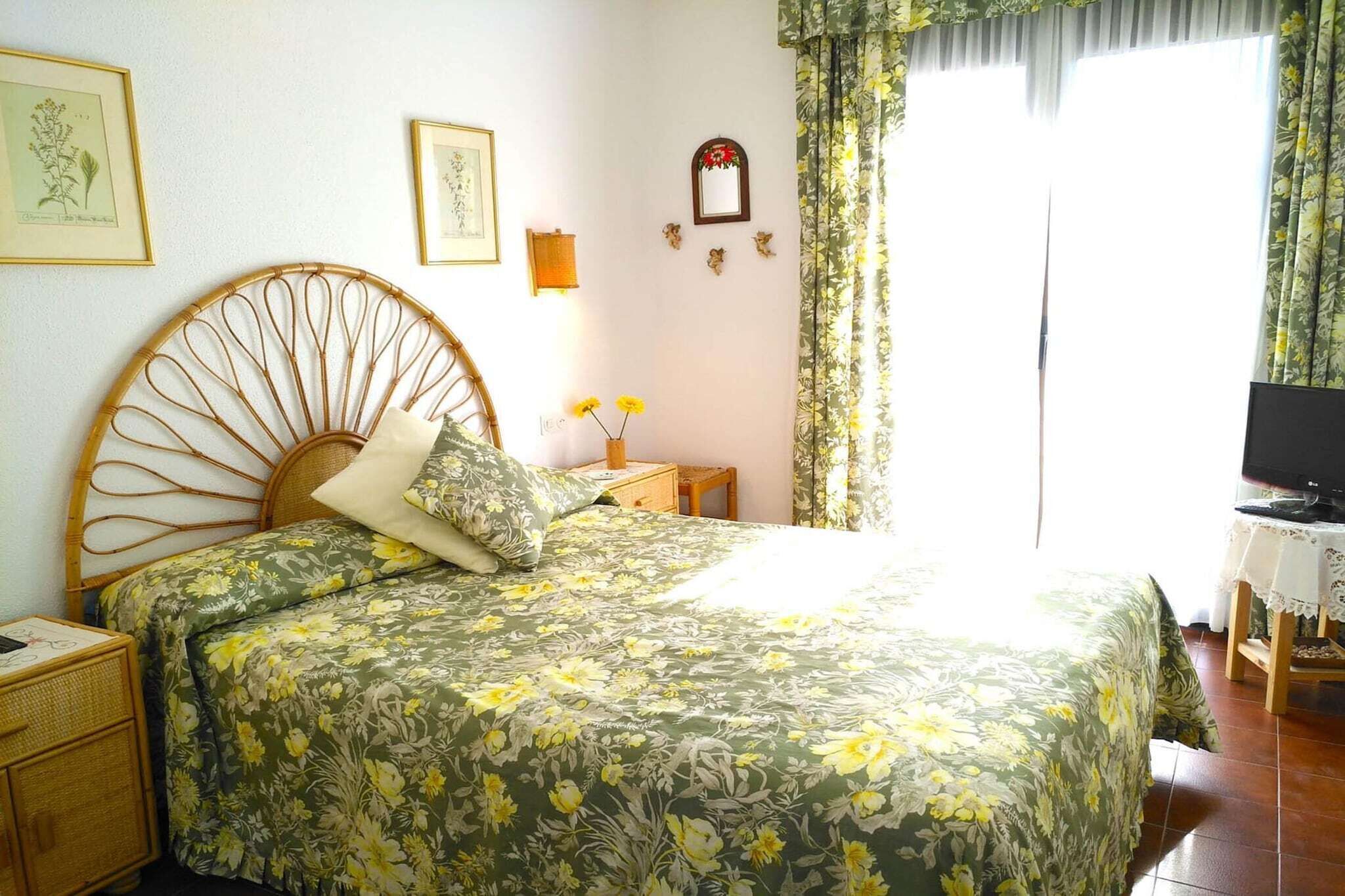 A bedroom with a bed, a nightstand, a bedside table with a lamp, a television, and a window with curtains.