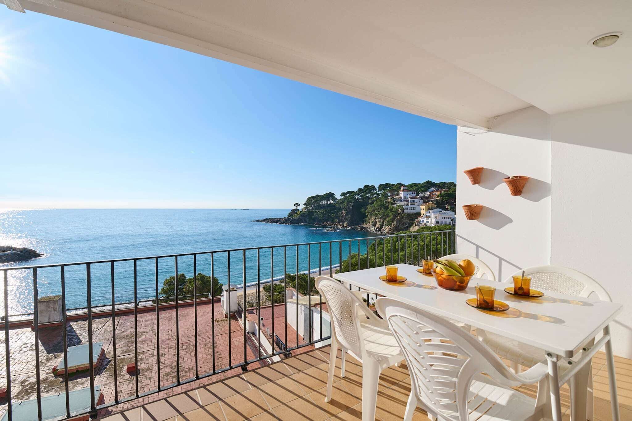 Un balcón con una mesa dispuesta para dos personas, con vistas a la costa, casas y follaje.