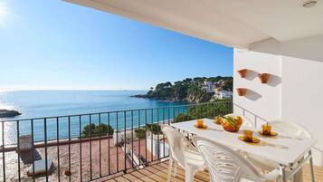 Un balcón con una mesa dispuesta para dos personas, con vistas a la costa, casas y follaje.