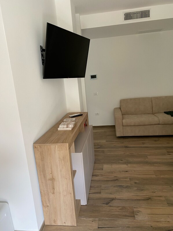 A living room with a flat-screen TV mounted on the wall, a wooden console table, a beige sofa, and a wooden floor.
