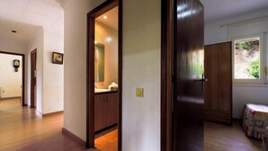 Um corredor com piso de madeira, uma porta que dá para um banheiro e uma pequena mesa com colcha.