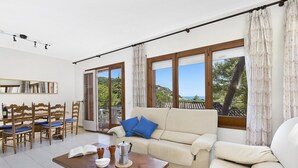 A living room with a sofa, chairs, a coffee table, and large windows offering a view of greenery and a building. - Apartment in Llafranc near Beach & Pool (Llafranc)