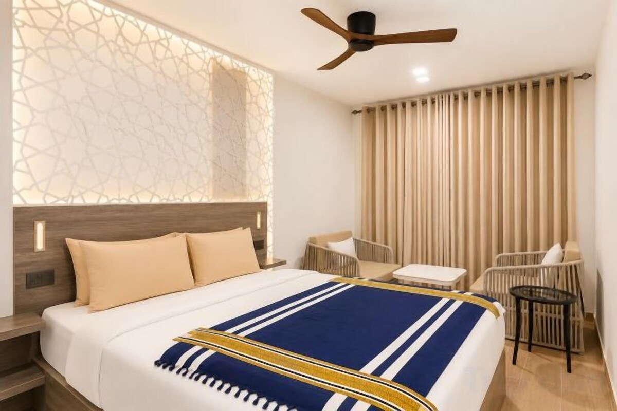 Habitación de hotel con cama, ventilador de techo, ventana con cortinas y una pequeña mesa con una silla.
