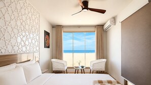 Une chambre d’hôtel avec un lit, deux chaises, une petite table, un ventilateur de plafond et une vue sur la plage.