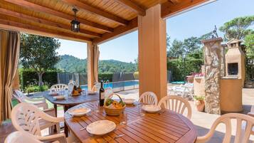 Un espace de restauration en plein air avec une table en bois et des chaises blanches, entouré d’un mur de pierre et offrant une vue sur un paysage montagneux.