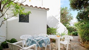 A patio with white plastic chairs and a table, surrounded by greenery and a white building with a tiled roof.