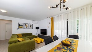 Ett modernt vardagsrum med en grön soffa, en gul pall, en platt-TV och ett matbord med en bordslöpare med blommönster.