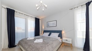 Ett modernt sovrum med en stor säng, ett sängbord, en liten avlastningsyta, en inramad tavla på väggen och utsikt genom fönstret.
