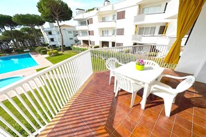Un balcon avec une table et des chaises blanches, une petite plante en pot et une vue sur une piscine et des immeubles d’habitation.