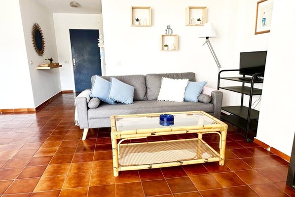 Un salon avec un canapé gris, des coussins bleus, une table basse en verre et un meuble TV noir.