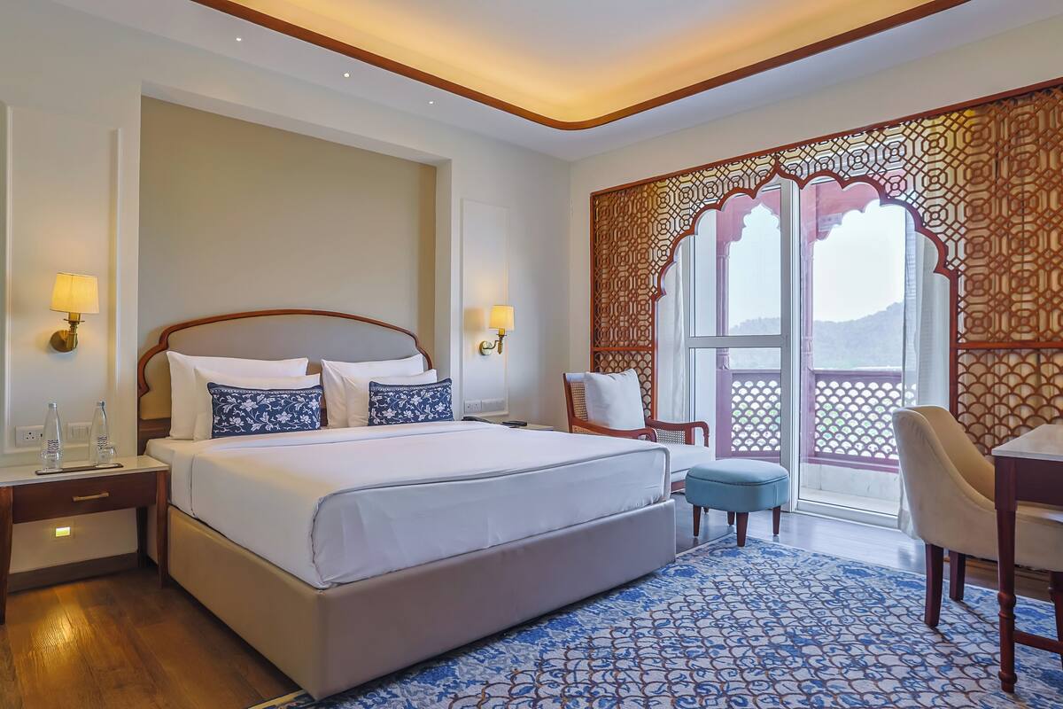 A hotel room with a large bed, bedside lamps, a desk, and a view through a decorative window.