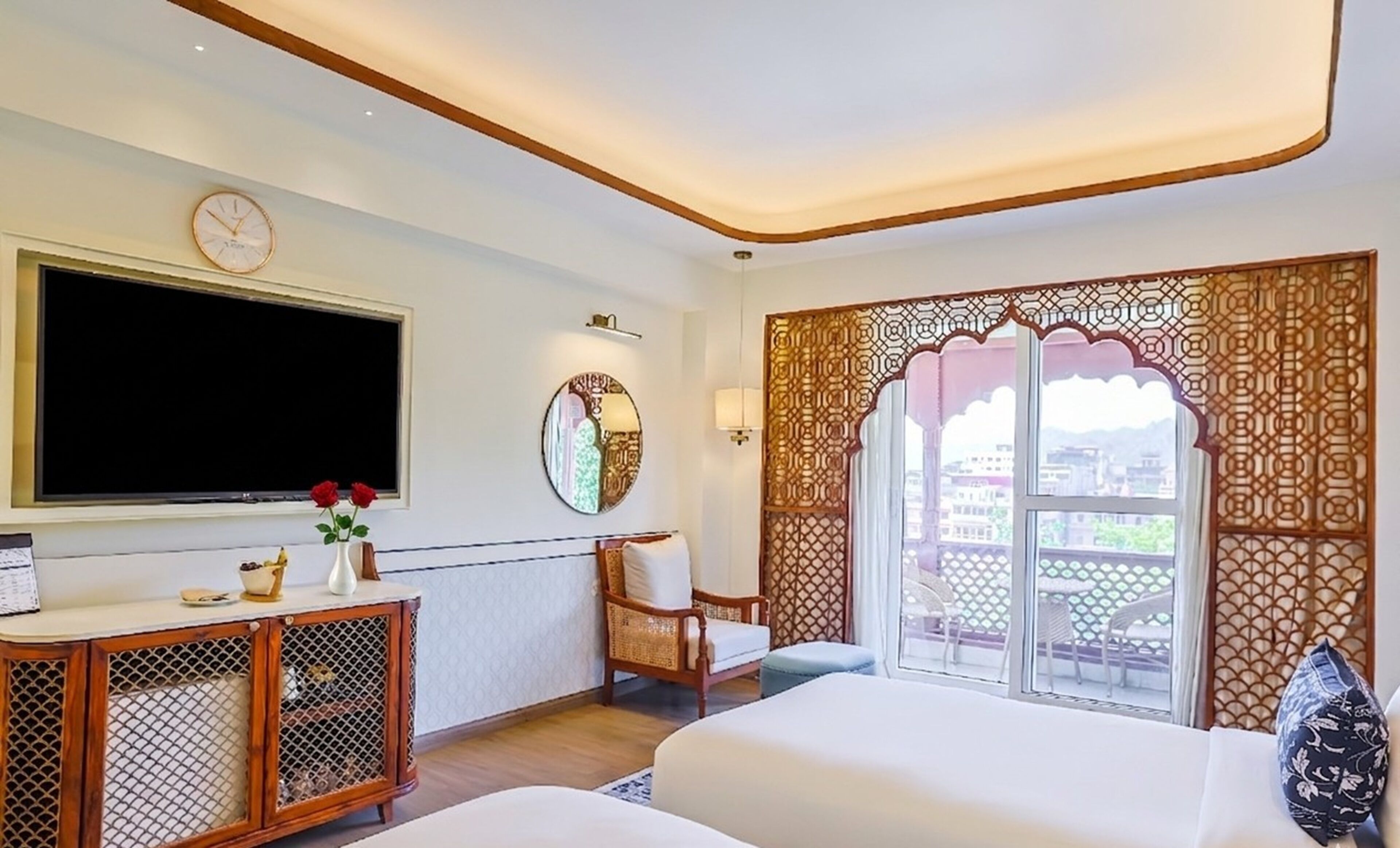 A hotel room with a large TV, a bed, a chair, a small table with a vase of flowers, and a decorative window view.
