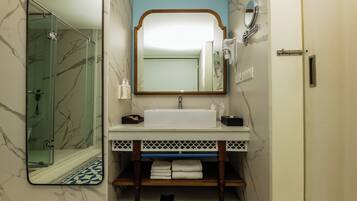 A bathroom with a glass shower enclosure, a white sink with a marble backsplash, and a mirror with a wooden frame.