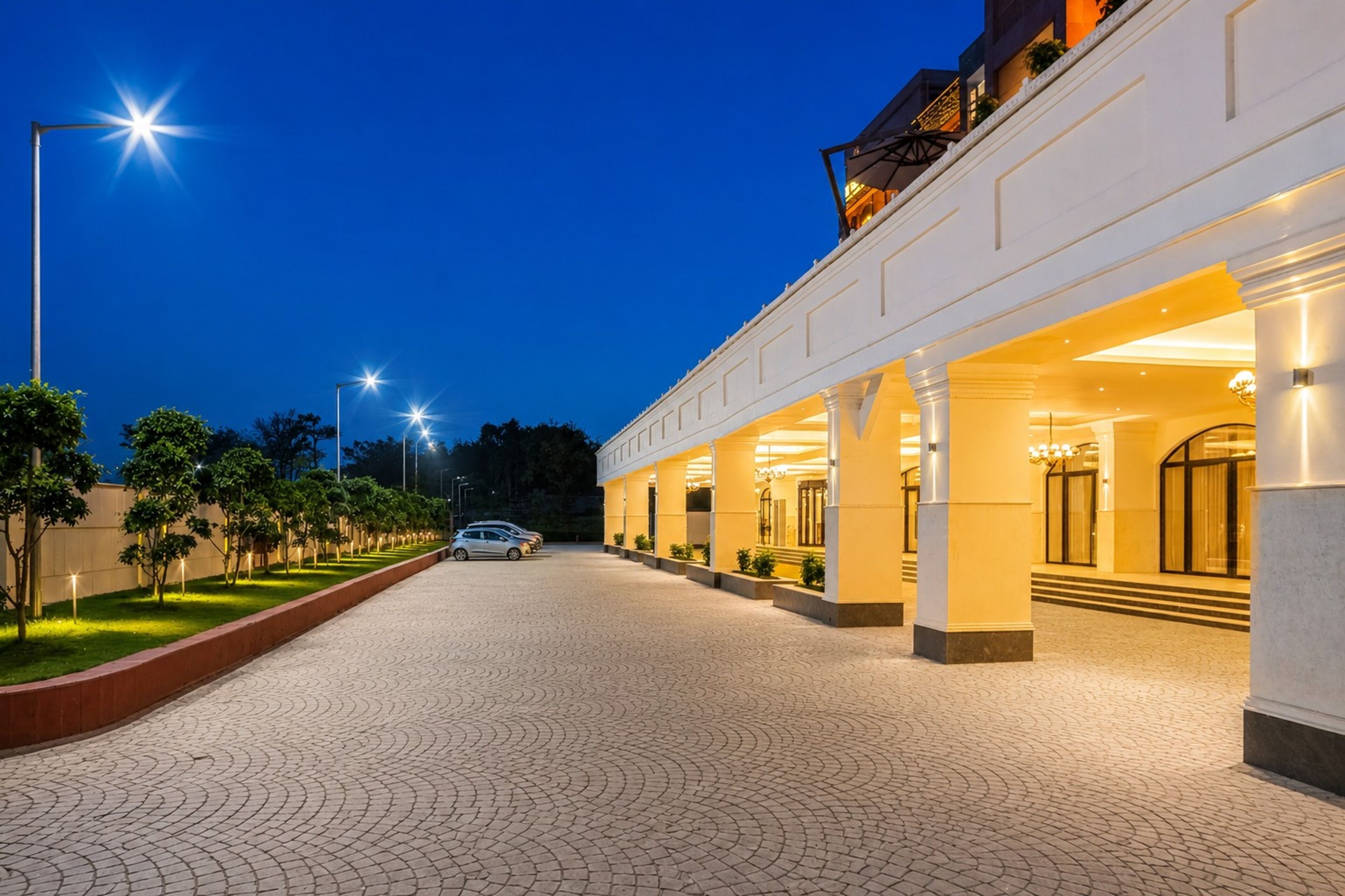 A hotel entrance with a paved driveway, a car parked in front, and a well-lit building facade.