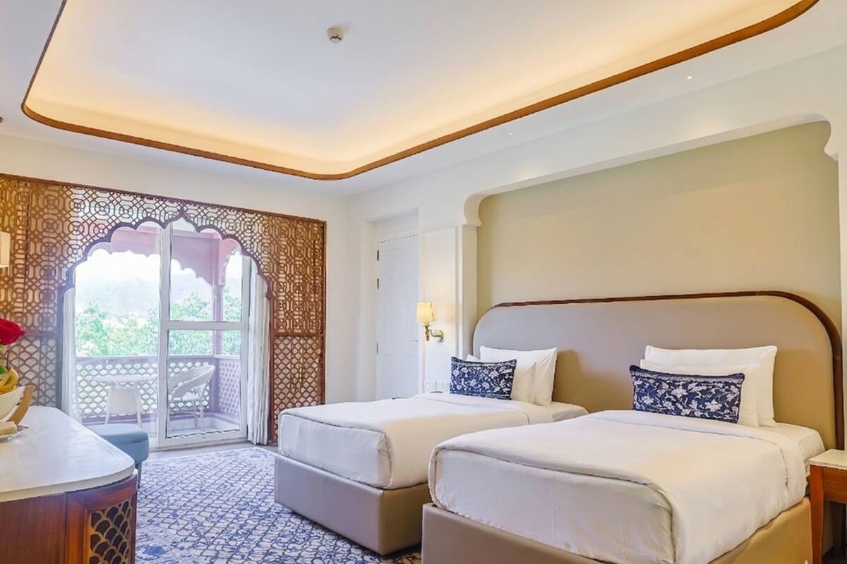 A hotel room with two beds, a sitting area with a table and a vase of flowers, and a view through a patterned door.