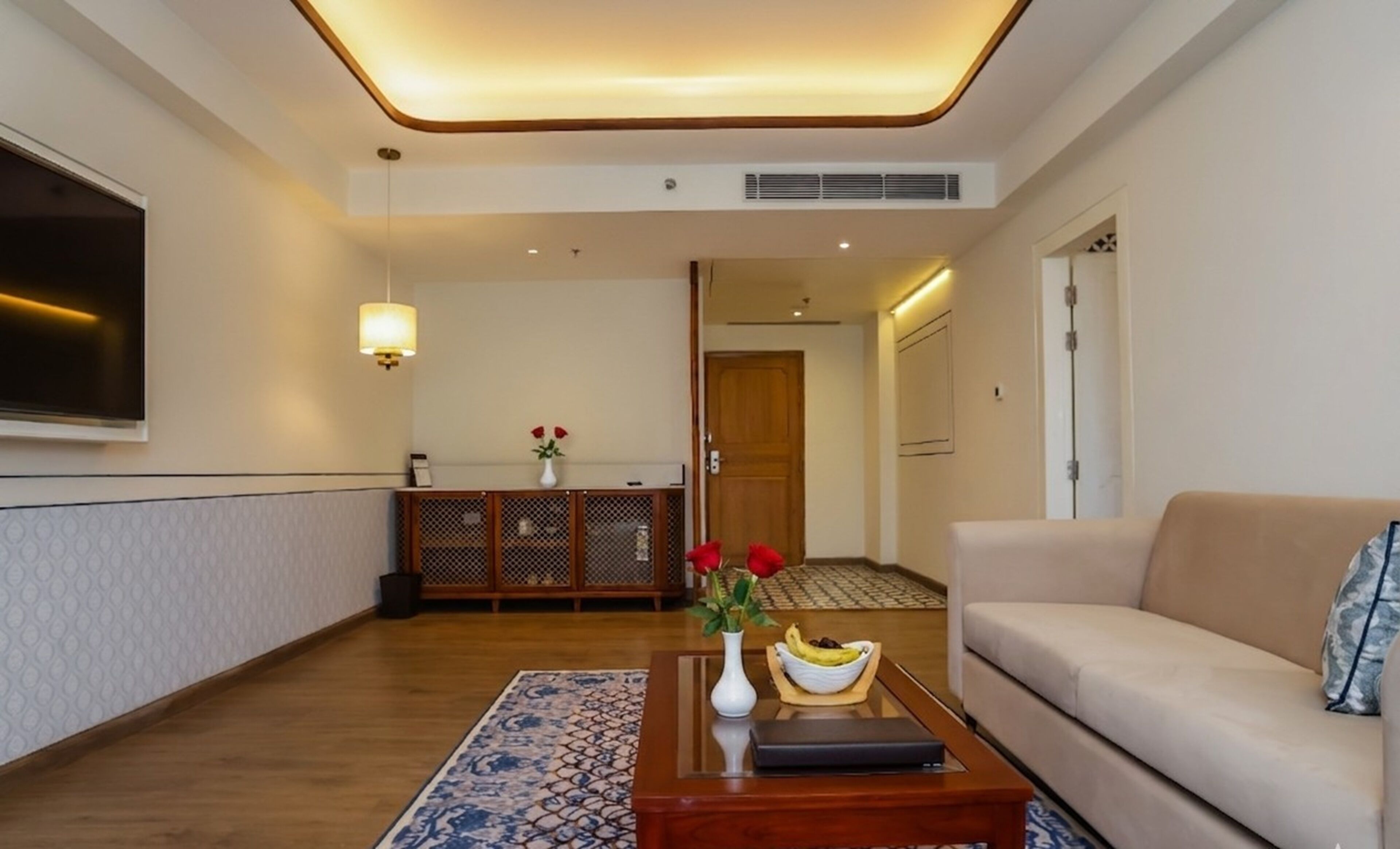 Um saguão de hotel com um sofá, uma mesa de centro com um vaso e uma tigela, uma televisão, um aparador decorativo e um tapete estampado.