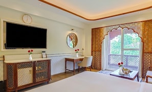 A hotel room with a large bed, a TV, a desk with a chair, a decorative wall piece, and a window with a view. - Bhagirathi By Atishay (Haridwar)