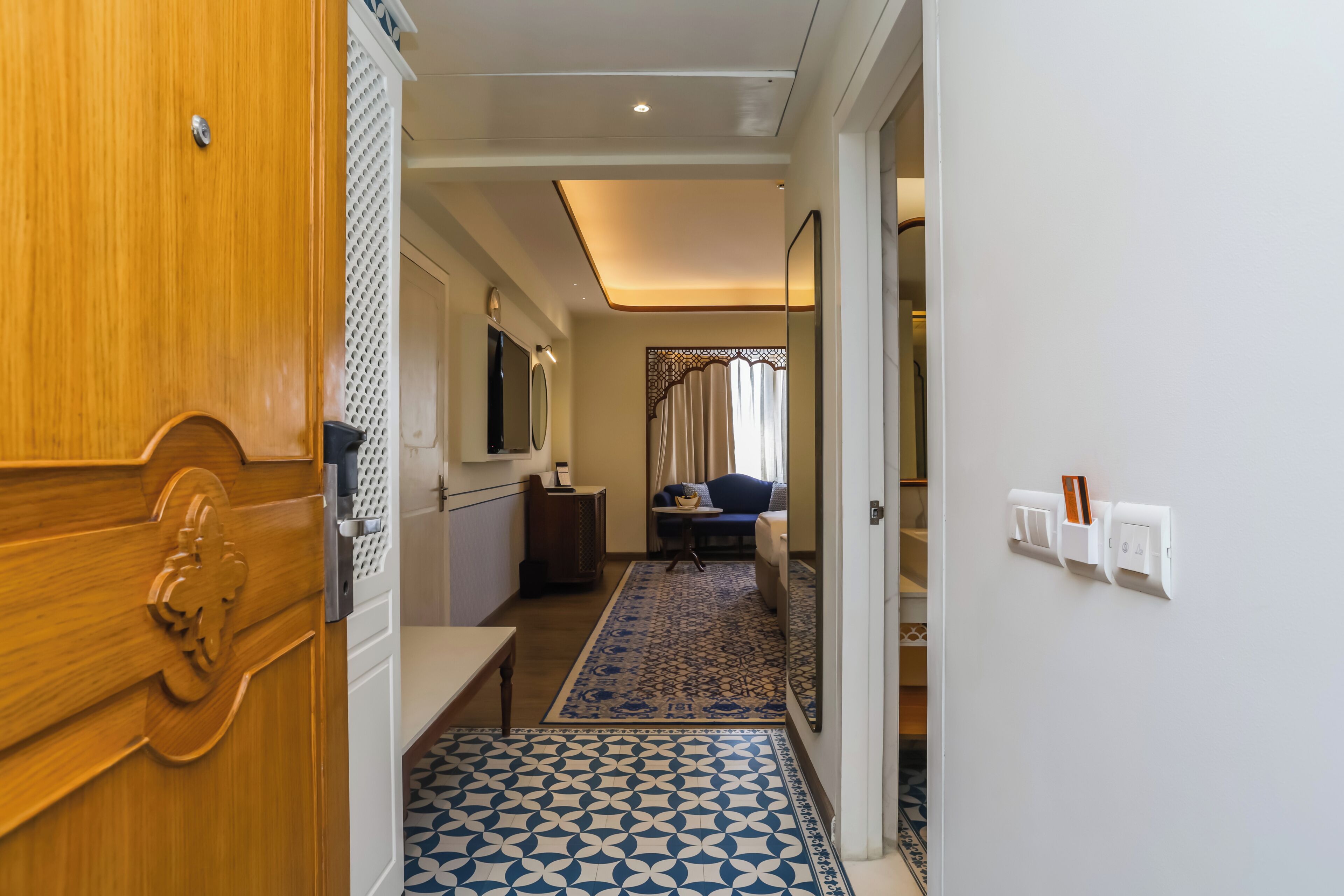 A hallway with a patterned floor, a wooden door, a mirror, a small table, and a seating area with a sofa and curtains.