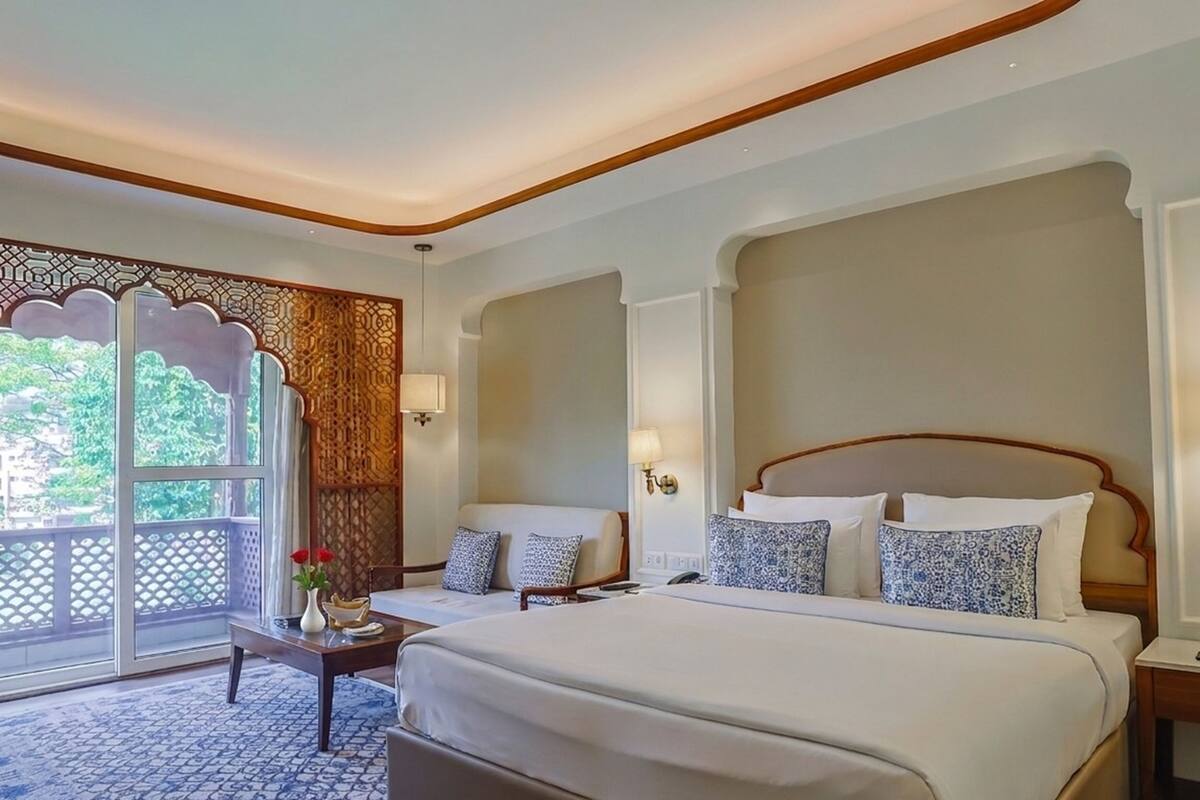 A hotel room with a large bed, two bedside tables, a small table with a vase and flowers, and a view through a window with a decorative screen.