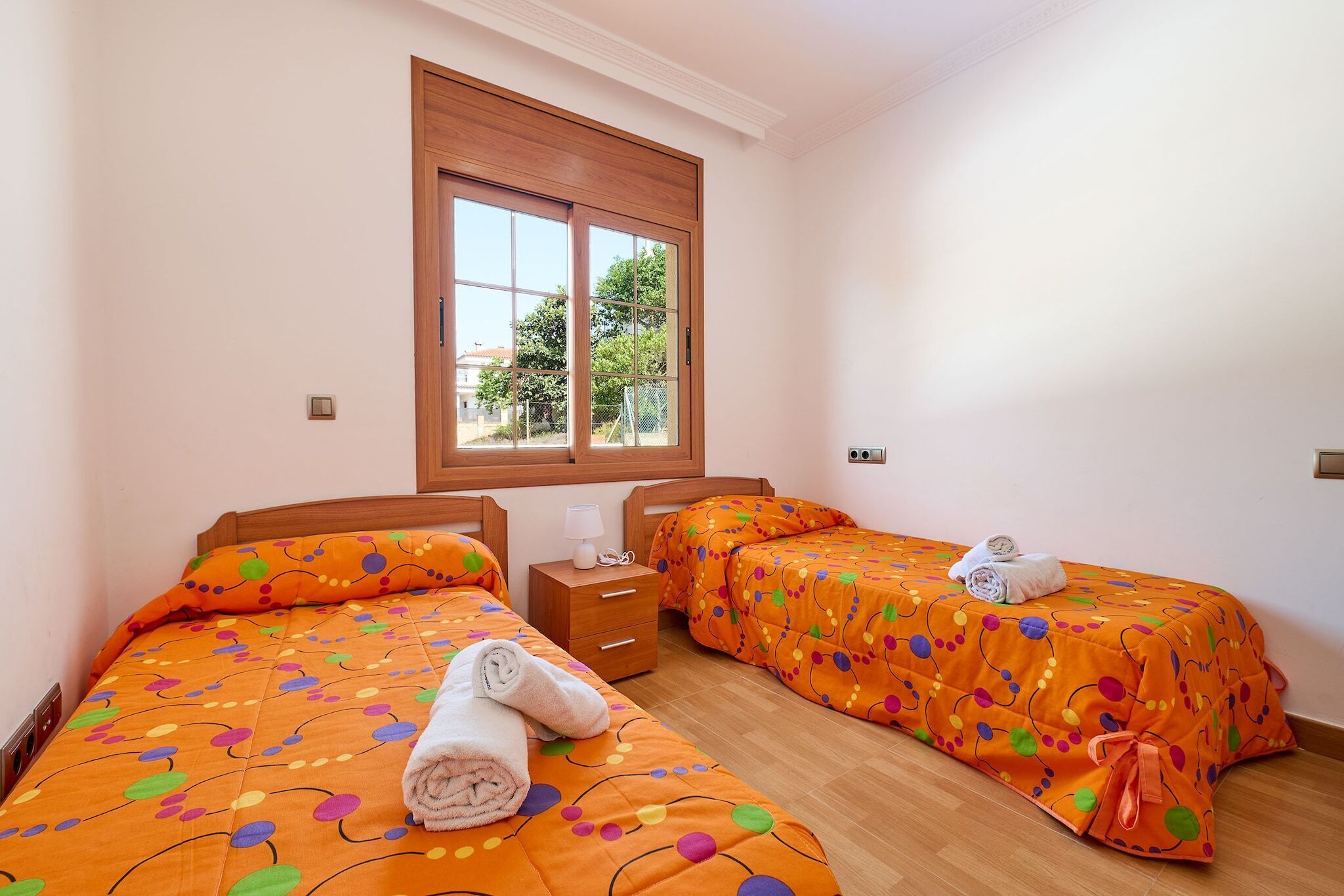 A room with two beds, each with an orange polka dot bedspread and matching pillows, a wooden headboard, a nightstand with a lamp, and a window with a view of trees and buildings.