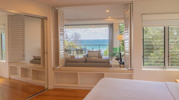 A bedroom with a large bed, a built-in window seat with a sofa and table, and a view of the ocean.